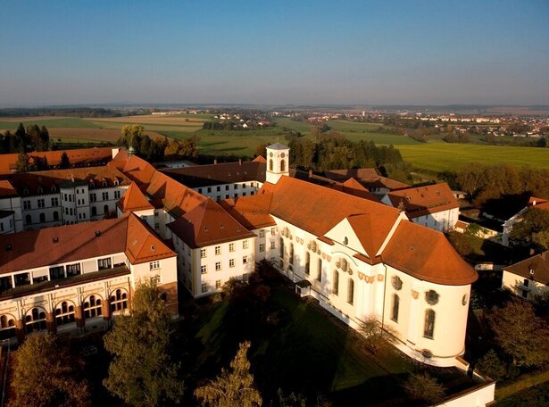 Luftaufnahme des Klosters Siessen in Oberschwaben bei Sonnenuntergang. Das historische Klostergebäude mit seinen weißen Wänden und roten Ziegeldächern liegt inmitten grüner Wiesen und weitläufiger Landschaften, die sich bis zum Horizont erstrecken.