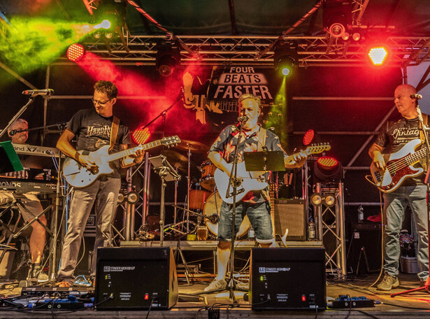 Rock- und Popband „Four Beats Faster“ live auf einer Open-Air-Bühne mit Gitarren, Bass, Keyboard und Schlagzeug, farbiges Bühnenlicht in Rot und Grün, energiegeladene Konzertatmosphäre bei Nacht.