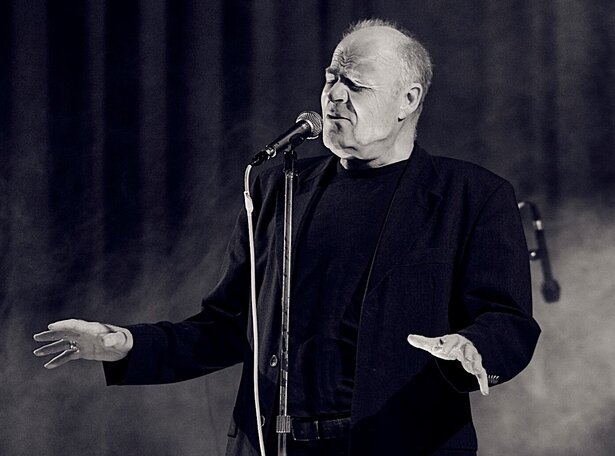 Der Sänger der Band Juice Cocker and Friends steht in dunklem Anzug vor Mikrofon mit ausgebreiteten Händen vor Schlagzeug mit Aufschrift 'Juice Cocker'. Ein Vorhang und die Beleuchtung deuten auf eine Bühnensituation hin. Das Bild ist in schwarz-weiß gehalten.