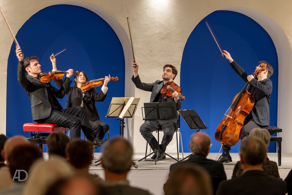 Klassik im Alten Kloster Streichquartett mit zwei Geigern, einem Bratschisten und einem Cellisten spielt vor Publikum in einem Raum mit weißen Rundbögen vor blauer Wand.