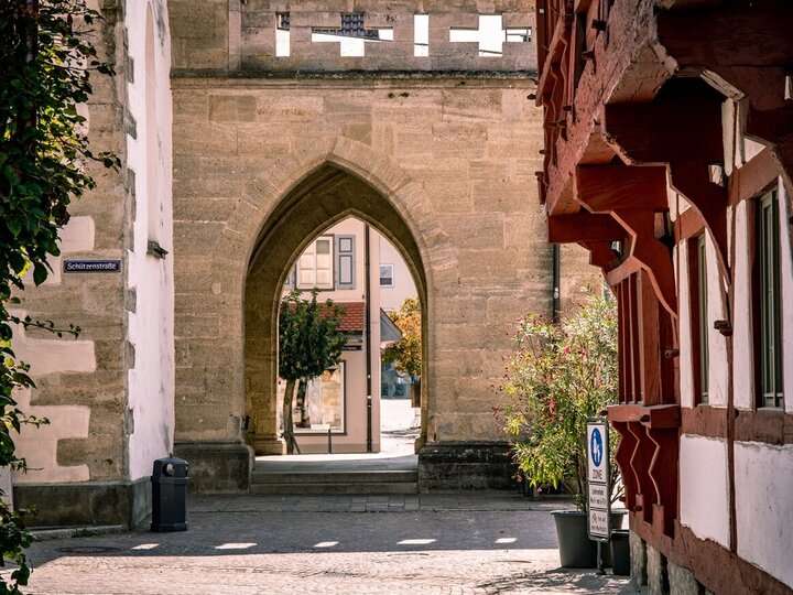 Stadtansicht eines historischen Kirchenportals mit Spitzbogen auf dem Marktplatz in Bad Saulgau, flankiert von Fachwerkhäusern und einer mit Pflanzen bewachsenen Mauer. Ein kleines Straßenschild mit der Aufschrift 'Schützenstraße' ist an der linken Wand angebracht, während im Hintergrund ein Baum und eine Häuserreihe zu sehen sind.