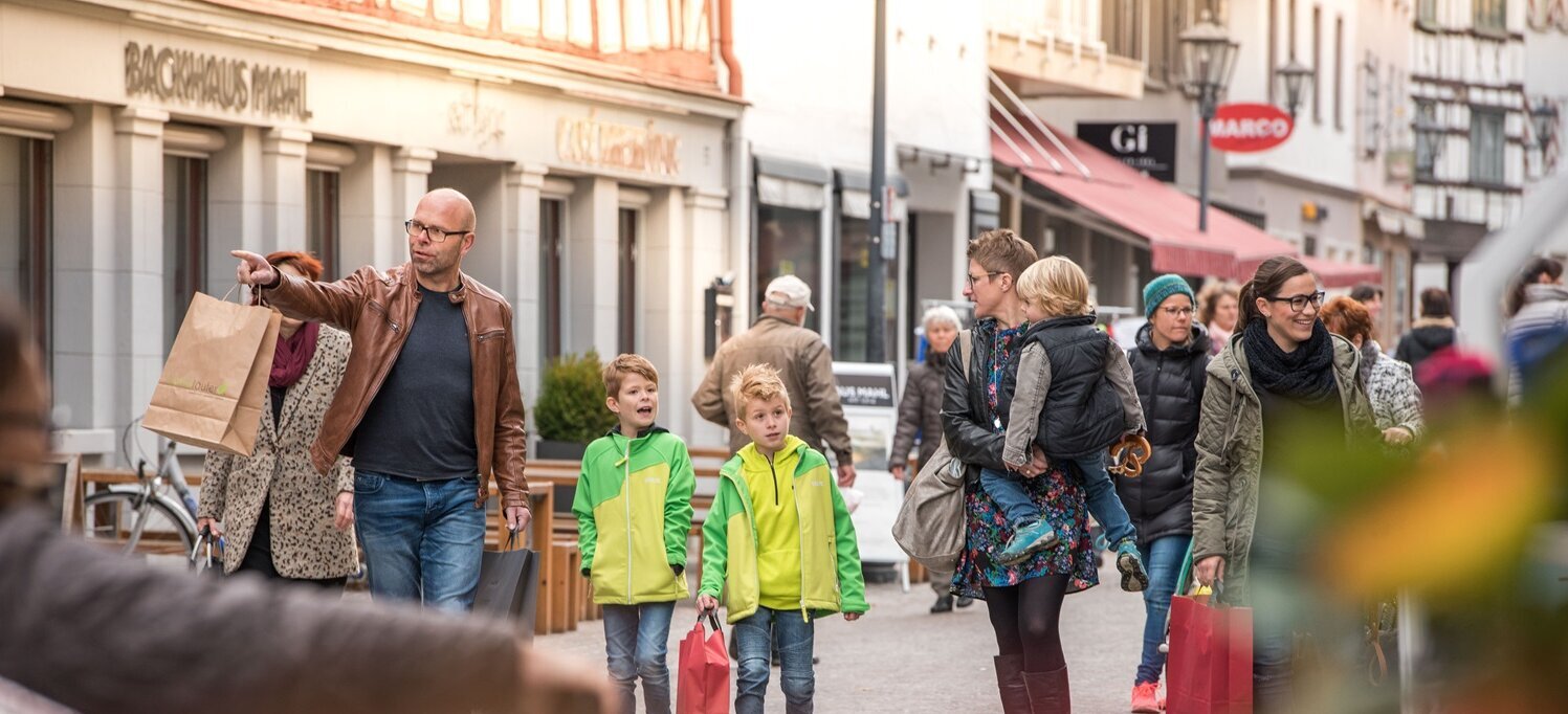 Eine Gruppe von Menschen in der belebten Fußgängerzone von Bad Saulgau; im Vordergrund ein Mann mit brauner Lederjacke, der mit Einkaufstaschen auf etwas zeigt, begleitet von zwei Jungen in leuchtend grünen Jacken. Eine Frau rechts trägt ein Kind auf dem Arm, während andere Personen mit Einkaufstaschen im Hintergrund zu sehen sind.