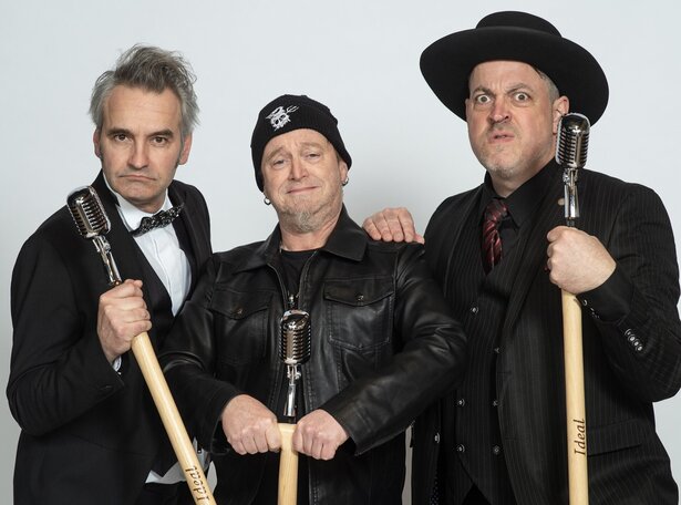 Drei mittelalte Männer in schwarzer Kleidung, teilweise mit schwarzer Kopfbedeckung, ziehen Grimassen und halten Mikrofonständer ins Bild. 