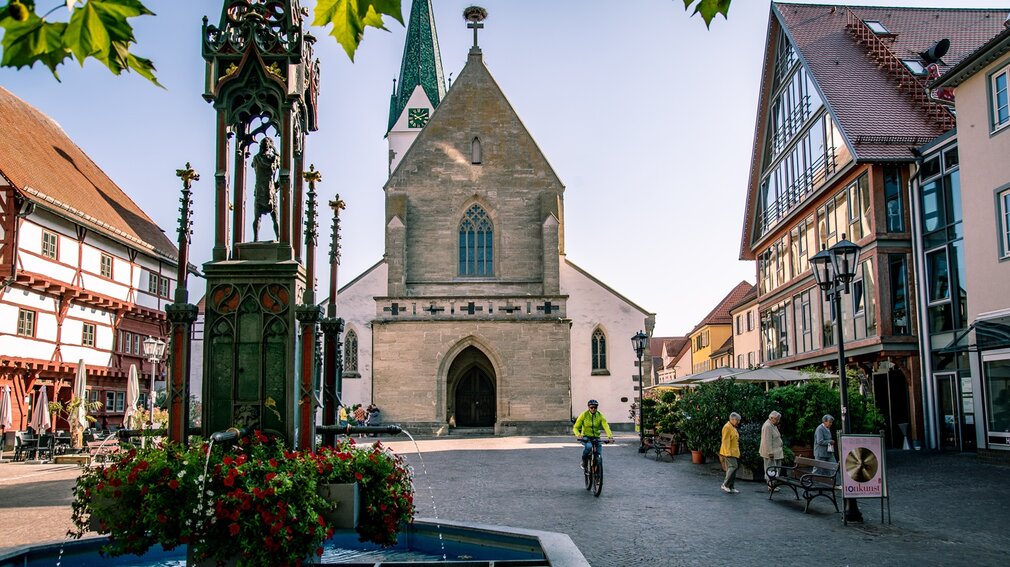 Historischer Marktplatz von Bad Saulgau mit gotischer Kirche, einem verzierten Brunnen mit Blumen sowie traditionellen und modernen Gebäuden; Menschen flanieren und ein Radfahrer fährt vorbei, bei sonnigem, Herbst-Wetter.
