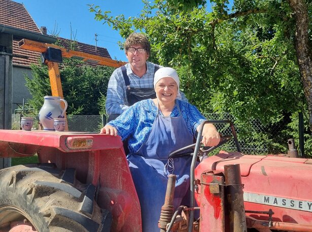 Zwei Personen auf einem roten Oldtimer-Traktor in ländlicher Umgebung, eine Frau sitzt lächelnd am Steuer, ein Mann steht hinter ihr, umgeben von Bäumen und einem Bauernhof