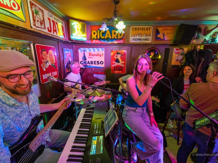 Liveband spielt in einer gemütlichen Bar: Sängerin mit Mikrofon steht im Mittelpunkt, umgeben von Bassist, Keyboarder, Schlagzeuger und weiteren Musiker:innen; bunte Lichter, dicht gedrängt stehendes Publikum und nostalgische Werbeschilder an den Wänden sorgen für Clubatmosphäre.