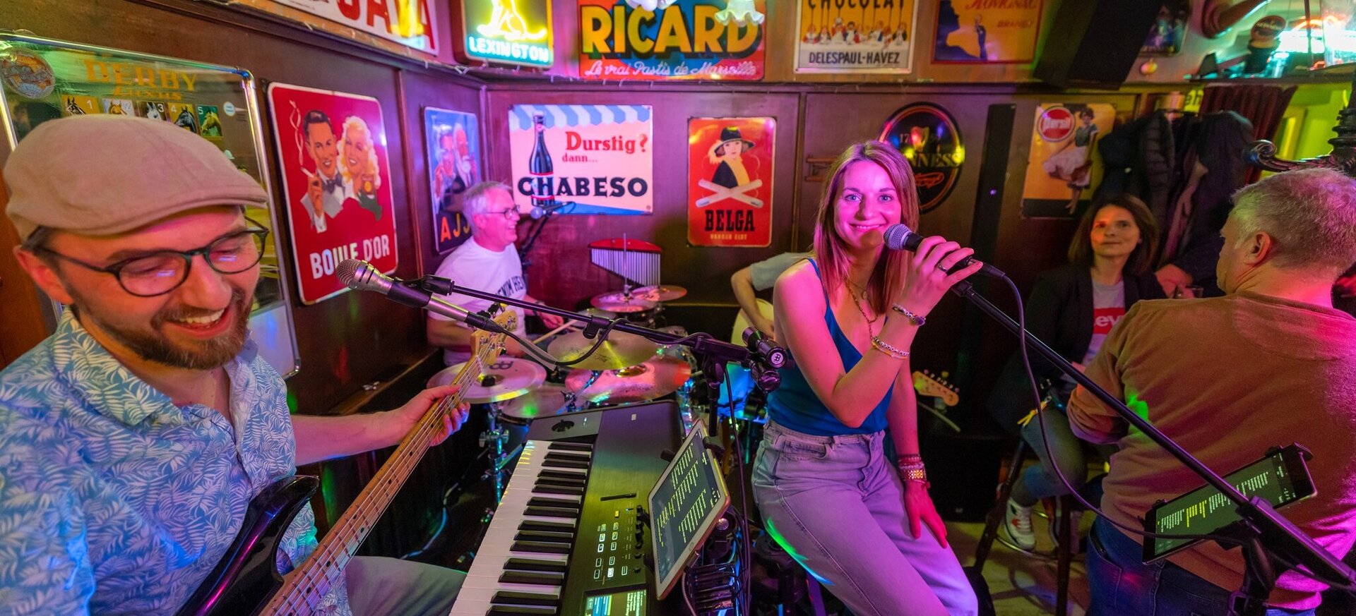 Liveband spielt in einer gemütlichen Bar: Sängerin mit Mikrofon steht im Mittelpunkt, umgeben von Bassist, Keyboarder, Schlagzeuger und weiteren Musiker:innen; bunte Lichter, dicht gedrängt stehendes Publikum und nostalgische Werbeschilder an den Wänden sorgen für Clubatmosphäre.