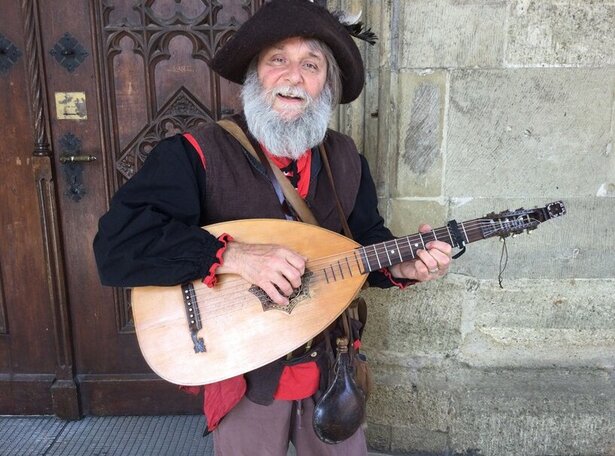 Mann in historischer Kleidung mit Dreispitz und Laute vor einer steinernen Wand und einem Holztor