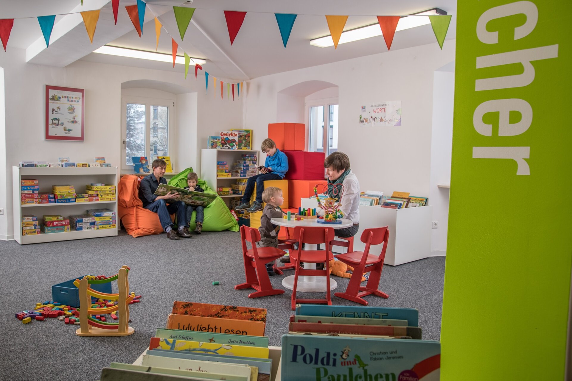 Kinder und Erwachsene spielen und lesen in einem bunten, gemütlichen Lese- und Spielbereich der Stadtbibliothek mit Sitzsäcken, Spielzeug und Bücherregalen