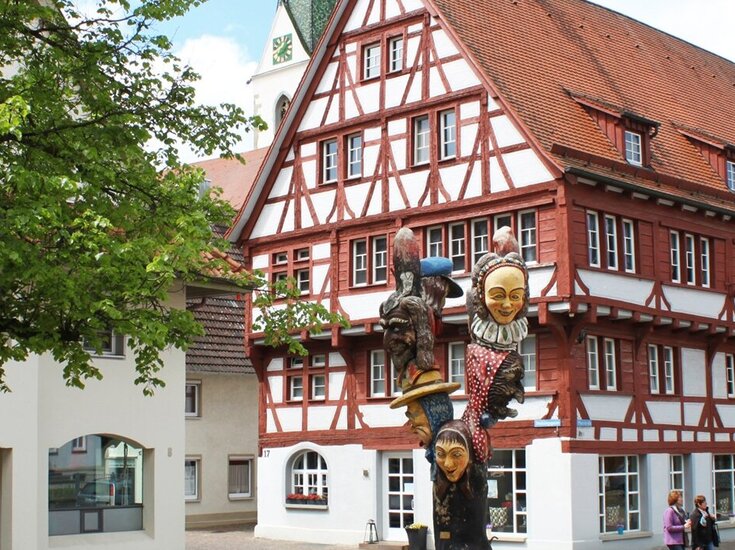 Der Narrenbrunnen mit bunten, maskierten Figuren in einem runden Becken vor einem Fachwerkhaus und einer Kirche im Hintergrund