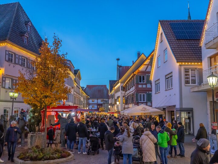 Stadt erleben Abendliche Szene auf dem belebten Nikolausmarkt in Bad Saulgau. Menschenmengen schlendern durch eine mit Lichterketten geschmückte Straße, umgeben von Fachwerkhäusern und moderner Architektur. Ein roter Verkaufsstand und ein Baum mit herbstlich gelben Blättern stehen im Vordergrund.