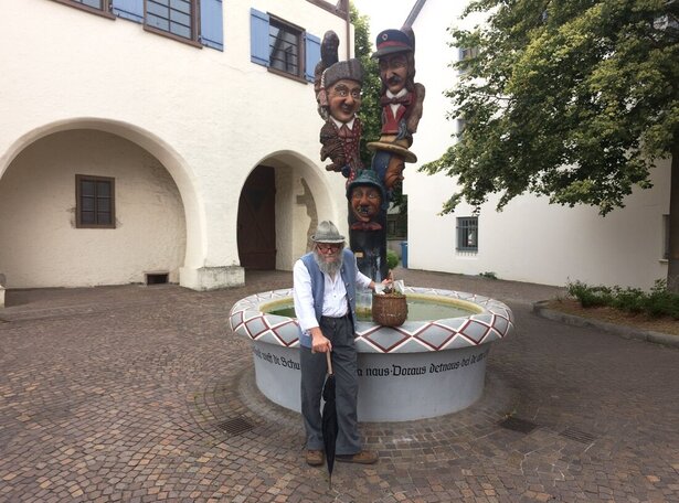 Älterer Mann mit Hut, Weste und Stock steht vor einem runden Brunnen mit bemaltem Rand und einer Säule mit mehreren bunten Figuren, umgeben von gepflastertem Platz und weißen Gebäuden mit Rundbögen