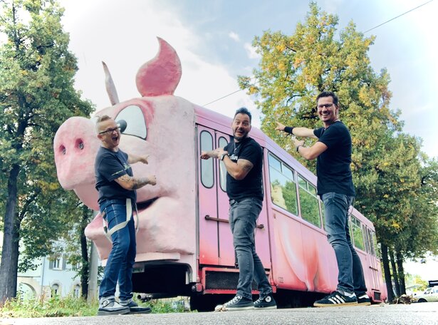 Drei Männer in schwarzen Band-Shirts stehen lachend vor einer pinken Straßenbahn in Form eines riesigen rosafarbenen Schweins und zeigen begeistert darauf; im Hintergrund stehen grüne Bäume an einer Straße.