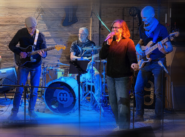 Liveband auf einer kleinen Bühne: zwei Sänger:innen mit Mikrofonen, Gitarrist, Bassist, Schlagzeuger und Keyboarder spielen gemeinsam vor einer Holzrückwand, beleuchtet in blau-orangem Bühnenlicht.