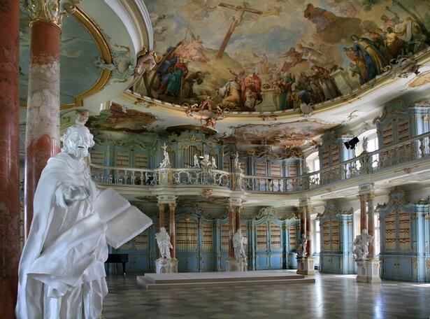 Prächtiger barocker Bibliothekssaal in Bad Schussenried mit kunstvoll bemalter Decke, verzierten Säulen und reich dekorierten blauen Bücherregalen. Eine weiße Statue steht im Vordergrund, während weitere Statuen entlang des Raums verteilt sind. Das Deckenfresko zeigt eine religiöse Szene mit zahlreichen Figuren und einem zentralen Kreuzmotiv, die dem Raum eine erhabene Atmosphäre verleiht.